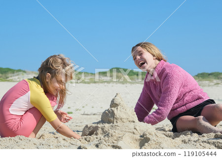 Two girls building a sandcastle on the beach under a clear blue sky. Two happy sisters having fun on the Atlantic ocean deserted beach. 119105444