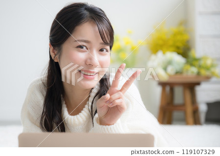 A young, cute woman in a white knitted sweater studying on a computer while lying face down, making a peace sign while looking at the camera, indicating her qualification or passing an exam. 119107329