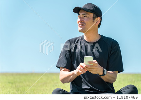 An Asian man in sportswear sitting on a yoga mat on the grass in a park and using a smartphone 119107469