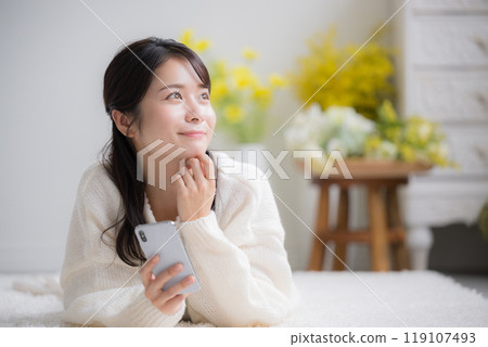 A young woman wearing a white knitted sweater lying face down and looking up while holding a smartphone A young woman wearing a white knitted sweater lying face down and looking up while holding a smartphone 119107493