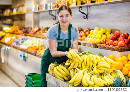 Female seller holding bananas standing in fruit and vegetable section of supermarket Female seller holding bananas standing in fruit and vegetable section of supermarket 119109582