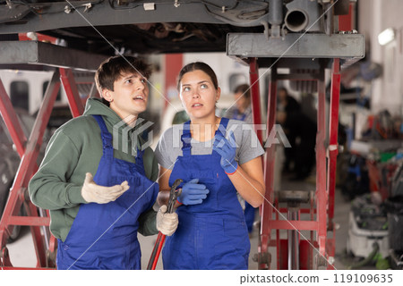 Two young mechanics discussing car repairs under lifted vehicle Two young mechanics discussing car repairs under lifted vehicle 119109635