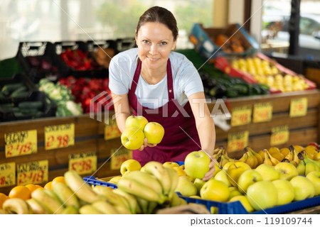 Female employee was distracted for minute and stands smiling with ripe apple in hands Female employee was distracted for minute and stands smiling with ripe apple in hands 119109744
