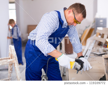 Professional builder in blue overalls cuts an aluminum profile with angle grinder 119109772