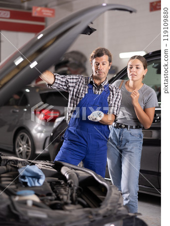 Woman talking to auto mechanic in uniform about repairing his car engine in auto repair shop 119109880