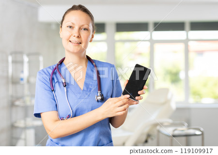 Woman doctor holds phone in hands and points finger at dark screen of gadget. 119109917