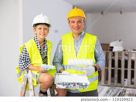 Portrait of father and son in yellow vest and protective helmet, who paint walls of room with paint roller Portrait of father and son in yellow vest and protective helmet, who paint walls of room with paint roller 119109923