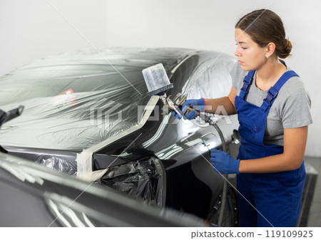Car painting and automobile repair service. Woman in blue overalls paints car with airbrush pulverizer in paint chamber Car painting and automobile repair service. Woman in blue overalls paints car with airbrush pulverizer in paint chamber 119109925