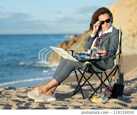 Businesswoman combining work with whiskey by sea on sunny day Businesswoman combining work with whiskey by sea on sunny day 119110050