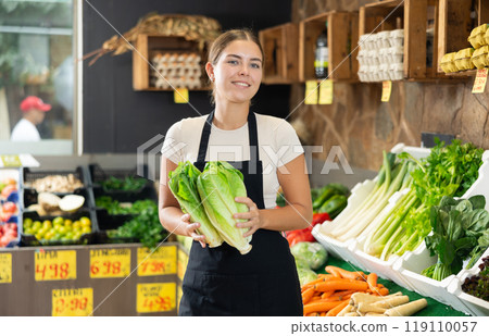 Young female seller in apron displaying assortment of lettuce buds at supermarket Young female seller in apron displaying assortment of lettuce buds at supermarket 119110057