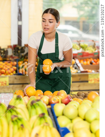 Interested salesgirl laying out oranges on display stand in supermarket Interested salesgirl laying out oranges on display stand in supermarket 119110217