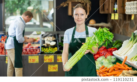 Woman seller of fruit and vegetable store works near showcase with garden-stuff, sorting celery Woman seller of fruit and vegetable store works near showcase with garden-stuff, sorting celery 119110231