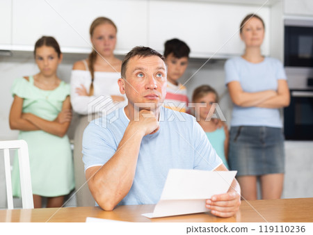Upset father reads and looks through financial documents. Sad wife with children in background Upset father reads and looks through financial documents. Sad wife with children in background 119110236