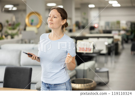 Woman wanders through furniture store with her mobile phone in hand Woman wanders through furniture store with her mobile phone in hand 119110324