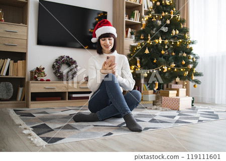 Young woman celebrating Christmas at home with phone and gifts Young woman celebrating Christmas at home with phone and gifts 119111601