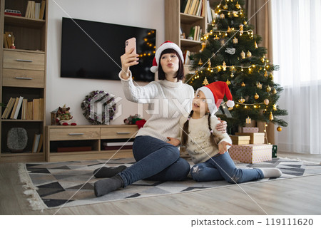 Mother and daughter taking selfie by Christmas tree at home Mother and daughter taking selfie by Christmas tree at home 119111620