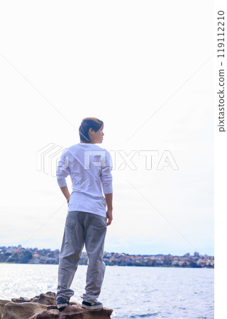 Back view of a Japanese man standing on a rock by the sea and gazing into the distance 119112210