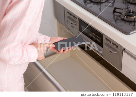 A middle-aged woman holding a smartphone while looking at the kitchen cabinet storage A middle-aged woman holding a smartphone while looking at the kitchen cabinet storage 119113157