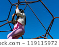 A toddler playing on a rope jungle gym, face not visible, against a clear, cloudless blue sky 119115972