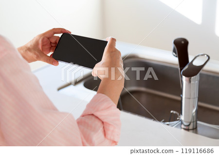 A middle-aged woman holding a smartphone and a kitchen sink on the counter A middle-aged woman holding a smartphone and a kitchen sink on the counter 119116686