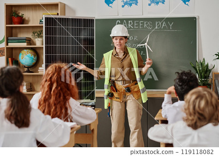 Female engineer holding wind turbine model standing by solar panel and telling kids about renewable energy sources Female engineer holding wind turbine model standing by solar panel and telling kids about renewable energy sources 119118082