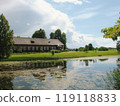 Scenic old house by a tranquil pond under a clear blue sky, surrounded by lush greenery, reflecting peaceful rural life. 119118833