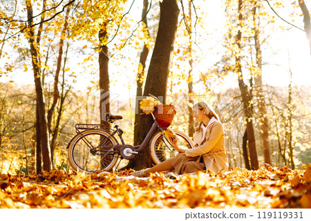 Beautiful young woman sitting on a fallen autumn leaves in a park, reading a book. Relaxation. 119119331