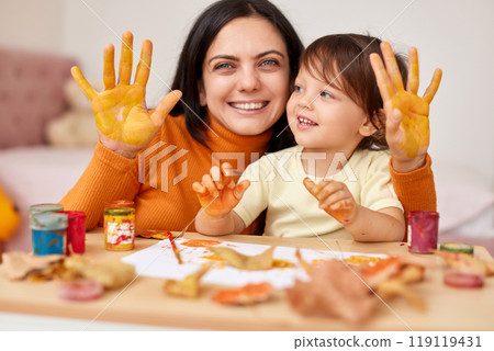 smiling little child girl and mother spending good time painting hands and autumn leaves together. mom and baby showing hands in yellow paint 119119431