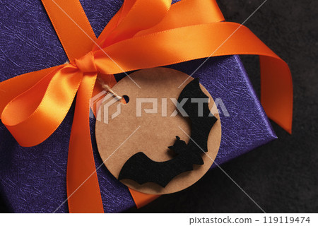 A Halloween gift wrapped in purple paper with orange ribbon and a round tag with a bat silhouette on it A Halloween gift wrapped in purple paper with orange ribbon and a round tag with a bat silhouette on it 119119474