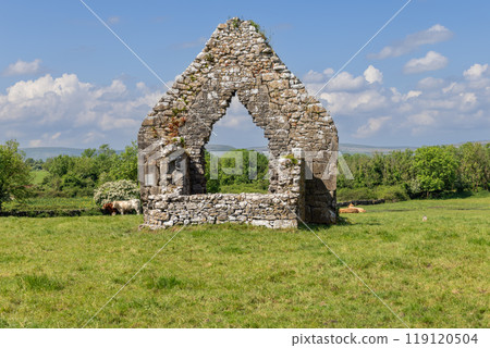 Weathered stone arch of Kilmacduagh Abbey in Galway stands against green fields and distant hills Weathered stone arch of Kilmacduagh Abbey in Galway stands against green fields and distant hills 119120504