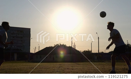 Silhouette of professional footballers kicking soccer ball with their heads on stadium at sunset. Two sportsmen showing tricks with ball while passing it to each other at field. Freestyle football 119121275
