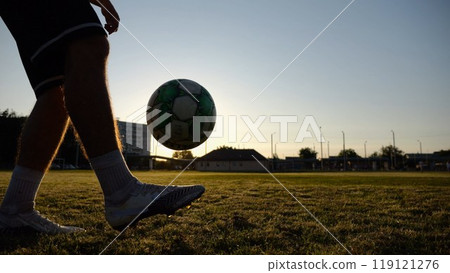 Legs of young man kicking ball at green field. Male feet of professional footballer juggling soccer ball on stadium at sunset. Sportsman practicing tricks outdoor. Concept of a freestyle football 119121276
