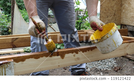 A construction worker in gray work clothes and gloves is making repairs and applying varnish or protective coating to a wooden structure. He is holding a brush and a bucket of varnish or paint. 119121422