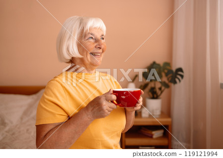 Caucasian blonde senior woman sitting on a white bed with a cup in her hand. Female relaxing and drinking cup of hot coffee or tea in the bedroom. 119121994