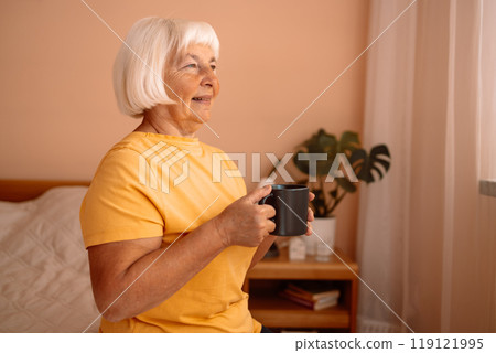 Caucasian blonde senior woman sitting on a white bed with a cup in her hand. Female relaxing and drinking cup of hot coffee or tea in the bedroom. 119121995