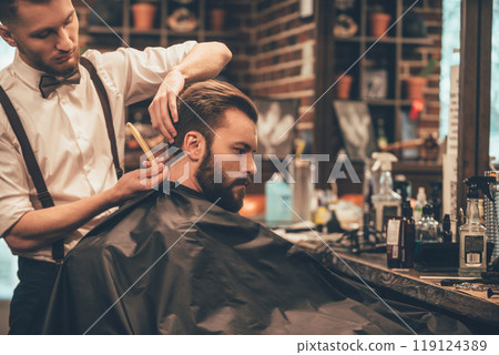 Making hair look magical. Young bearded man getting haircut with straight edge razor by hairdresser while sitting in chair at barbershop 119124389