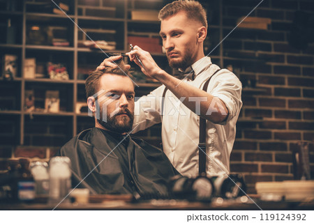 Making haircut look perfect. Young bearded man getting haircut by hairdresser while sitting in chair at barbershop Making haircut look perfect. Young bearded man getting haircut by hairdresser while sitting in chair at barbershop 119124392