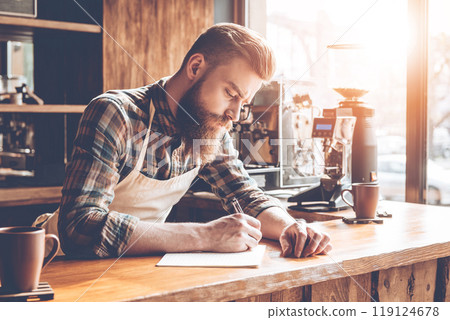Writing down new coffee recipes. Young bearded man in apron writing in notebook while leaning to bar counter at cafe Writing down new coffee recipes. Young bearded man in apron writing in notebook while leaning to bar counter at cafe 119124678