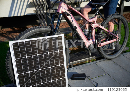 Pink electric mountain bike parked beside staircase, connected to small solar panel for charging. Bike rugged tires, situated on paved pathway with grassy lawn. Pink electric mountain bike parked beside staircase, connected to small solar panel for charging. Bike rugged tires, situated on paved pathway with grassy lawn. 119125065