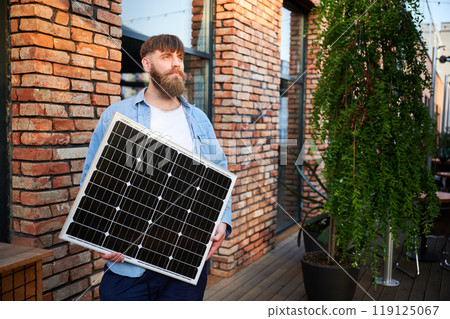 Portrait of bearded man holds solar panel, standing in front of brick building and lush greenery. Guy in shirt and t-shirt exudes calm and eco-conscious vibe, blending nature with urban elements. Portrait of bearded man holds solar panel, standing in front of brick building and lush greenery. Guy in shirt and t-shirt exudes calm and eco-conscious vibe, blending nature with urban elements. 119125067