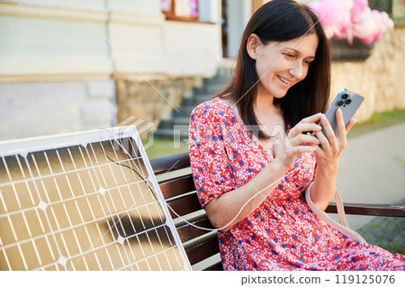 Happy women using smartphone connected to solar panel, smiling. Integration of sustainable renewable energy into everyday life, demonstrating practical use of solar power for charging devices. 119125076