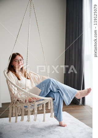 Young girl chilling at home in comfortable hanging chair near window. Gorgeous girl sitting on swing and smiling to camera. Pretty female in casual clothes relaxing in hanging chair indoor. Young girl chilling at home in comfortable hanging chair near window. Gorgeous girl sitting on swing and smiling to camera. Pretty female in casual clothes relaxing in hanging chair indoor. 119125079