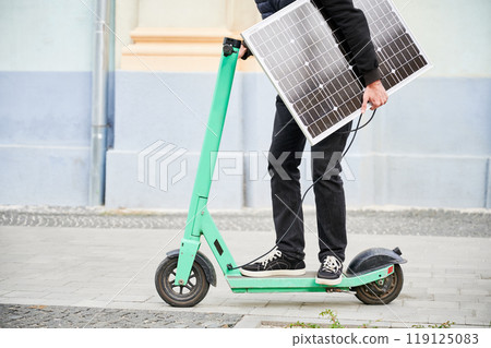 Cropped view of man rides electric scooter while carrying solar panel. Integration of solar power as sustainable energy source for charging electric scooters, promoting eco-friendly transportation. Cropped view of man rides electric scooter while carrying solar panel. Integration of solar power as sustainable energy source for charging electric scooters, promoting eco-friendly transportation. 119125083