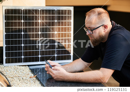 Bearded man using smartphone next to photovoltaic solar panel. Integration of sustainable renewable energy into everyday life, demonstrating practical use of solar power for charging devices. 119125131