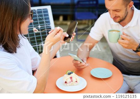 Man and woman charging smartphone from solar panel, relaxing in cafe. Cute couple using portable solar charger for devices. Charming couple sitting on terrace on background of solar battery. 119125135