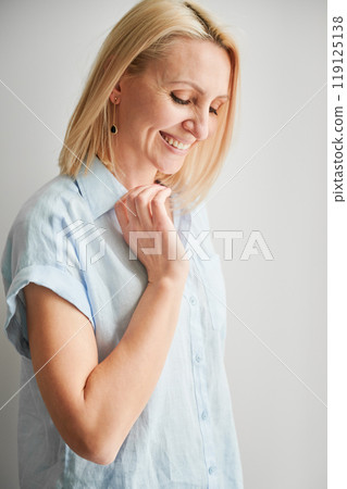 Gentle woman smiling and looking down. Close up portrait of pretty woman in blue shirt on light background. Gentle woman smiling and looking down. Close up portrait of pretty woman in blue shirt on light background. 119125138