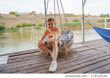 Middle-aged man sitting in wicker swing on terrace on beach. Middle-aged man sitting in wicker swing on terrace on beach. 119126146