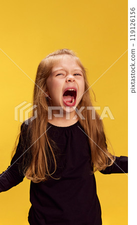 Little girl lets out loud, powerful yell, showing her temper against yellow studio background. Raw expression of anger or frustration. 119126156