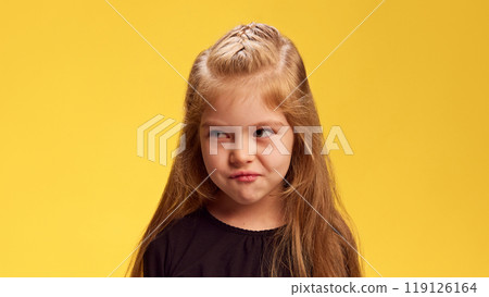 With mischievous expression, girl raises her eyebrows and looks sideways, almost plotting something playful against yellow studio background. 119126164