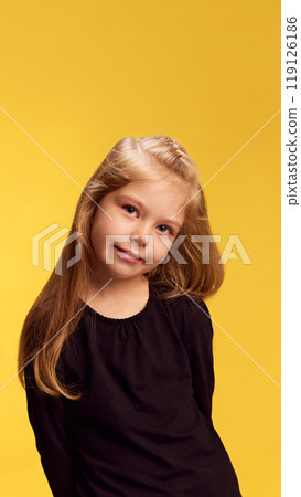 With soft, sweet expression, little girl tilts her head slightly to side, radiating innocence and charm against yellow studio background. 119126186